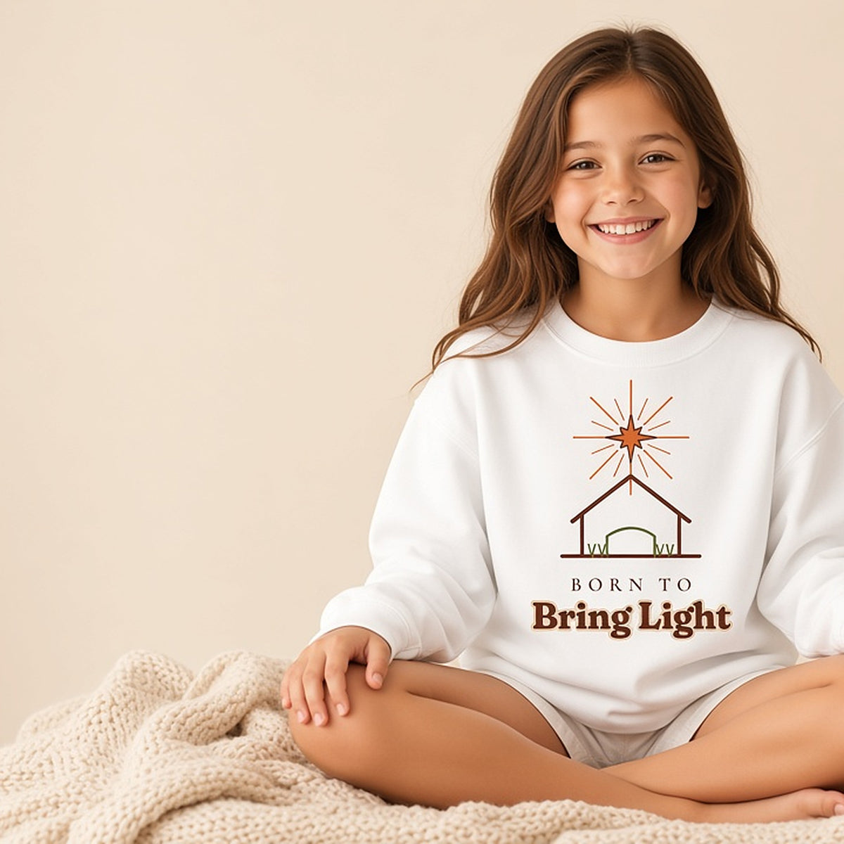 Child sitting in the white Born to Bring Light kids Christmas sweatshirt in a warm studio scene