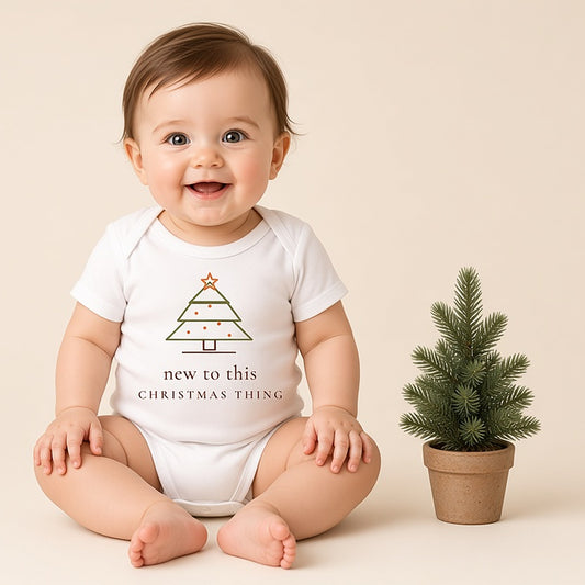 Baby sitting beside a mini Christmas tree in the New to This Christmas Thing bodysuit