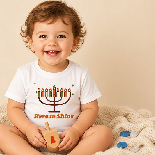 Toddler holding a wooden dreidel in the Here to Shine menorah tee