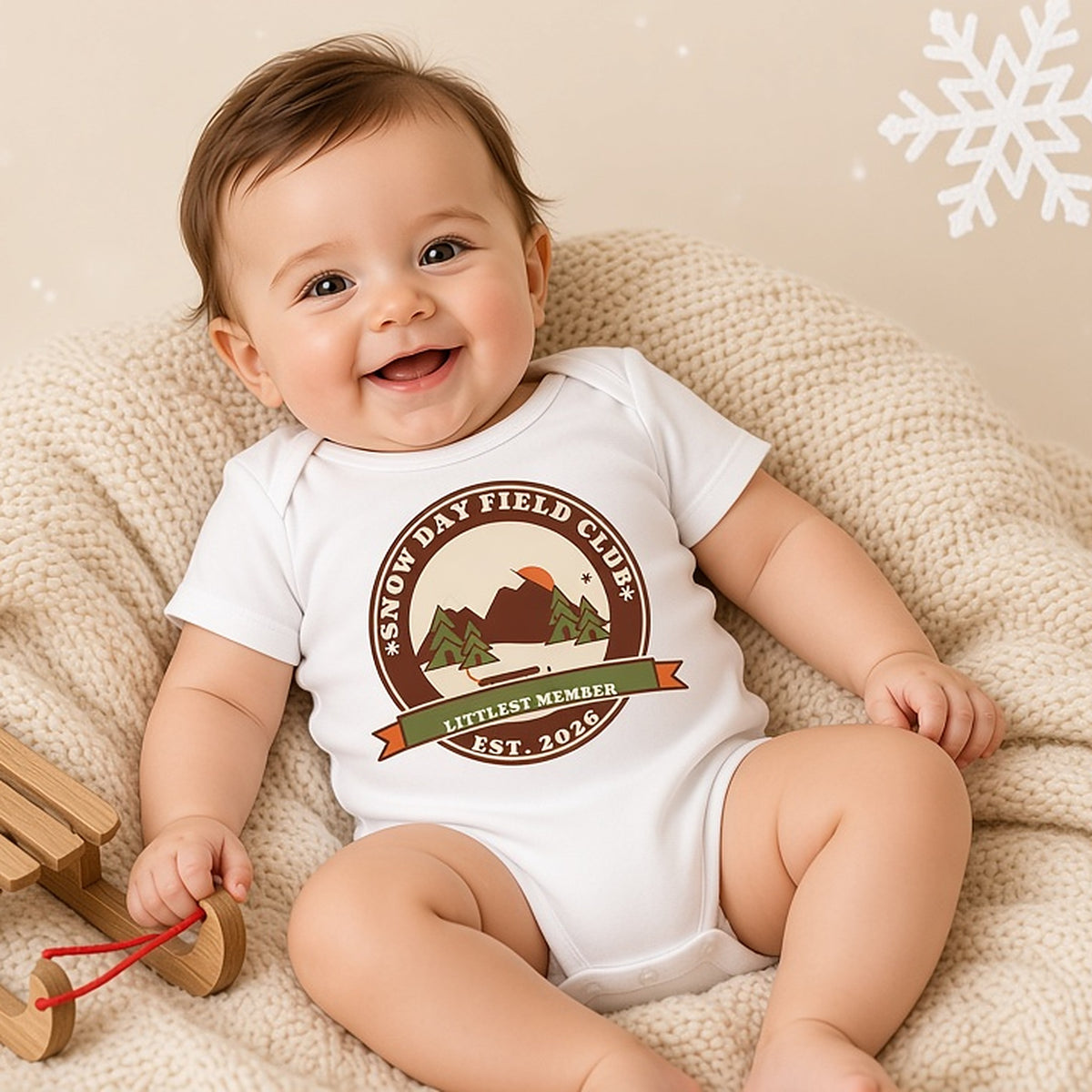 Baby lying on a cream blanket next to a toy sled in the Snow Day Field Club Littlest Member bodysuit