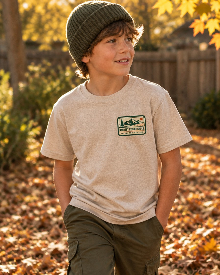 Kid wearing the Heather Dust Harvest Expedition Co. kids Thanksgiving tee with a mountain and pine trail badge