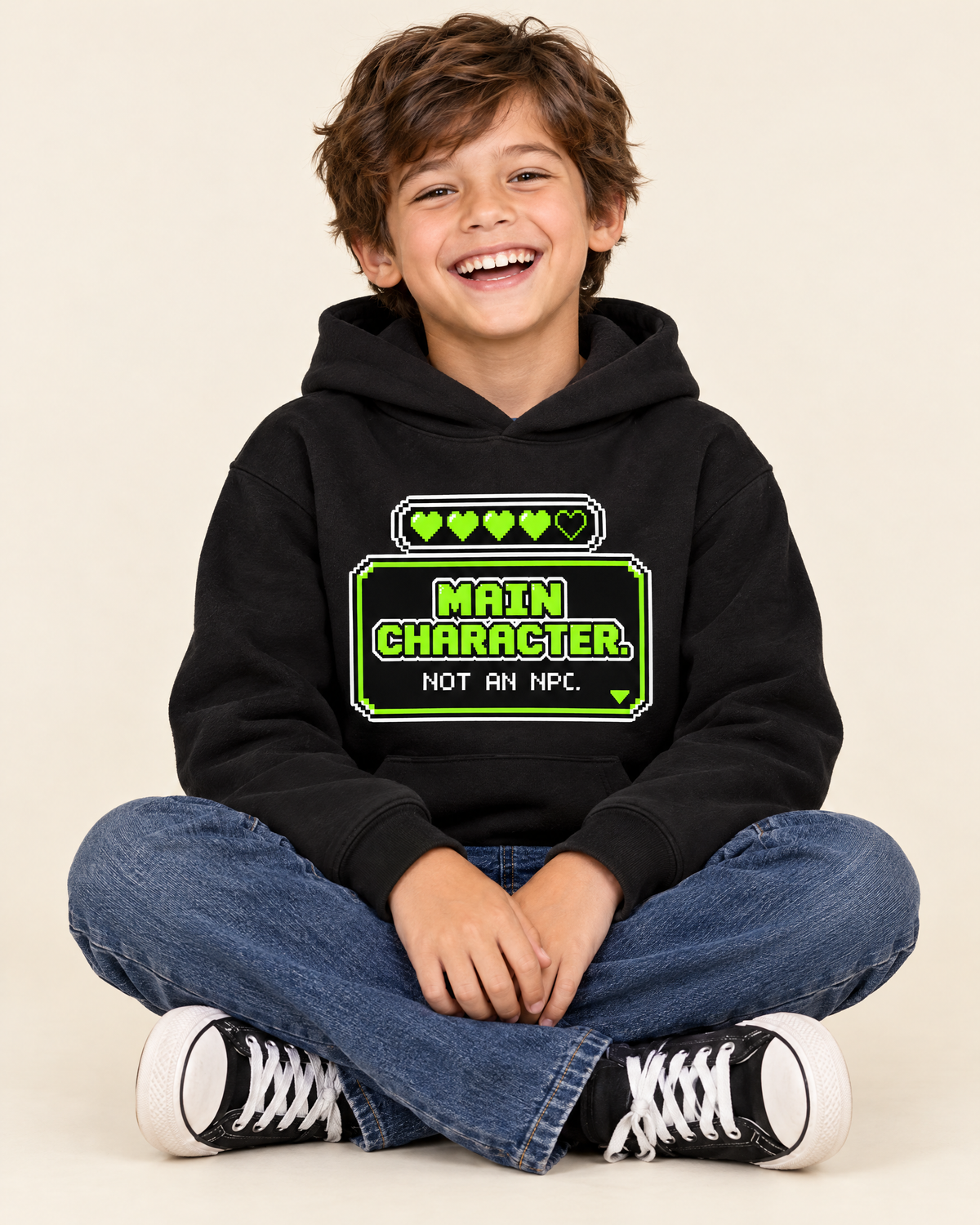 Laughing kid sitting cross-legged in the Main Character gamer hoodie
