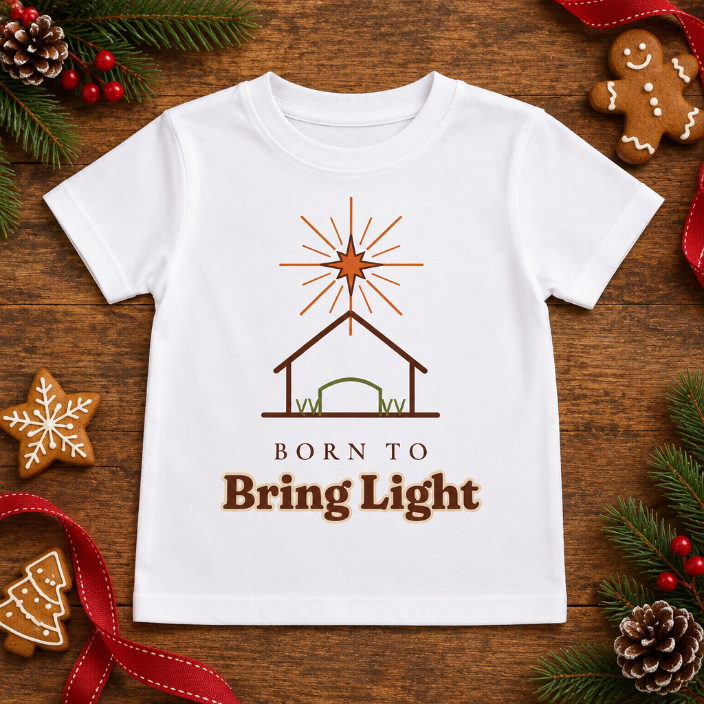 Flat lay of the Born to Bring Light Toddler Christmas Shirt: Faith Tee on a wooden table with pine sprigs and gingerbread cookies