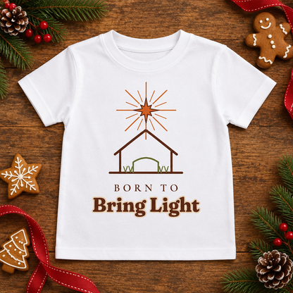 Flat lay of the Born to Bring Light Toddler Christmas Shirt: Faith Tee on a wooden table with pine sprigs and gingerbread cookies