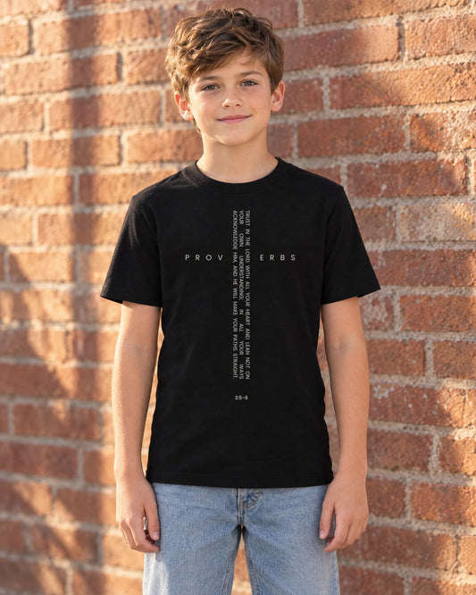 Boy wearing the black Proverbs Cross kids Christian t-shirt with the Proverbs 3:5-6 verse cross, standing by a brick wall