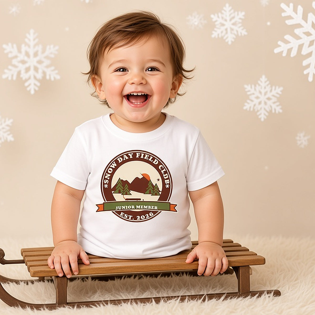Laughing toddler on a wooden sled in the Snow Day Field Club Junior Member tee