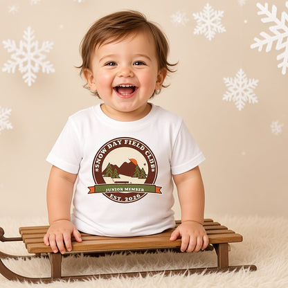 Laughing toddler on a wooden sled in the Snow Day Field Club Junior Member tee