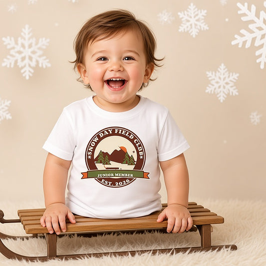 Laughing toddler on a wooden sled in the Snow Day Field Club Junior Member tee